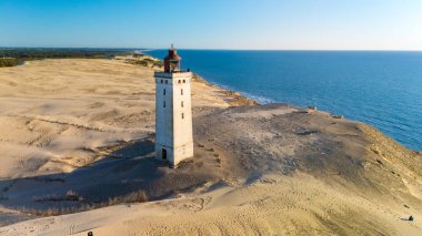 Rubjerg Knude deniz fenerinin çarpıcı manzarası, Danimarka kıyıları boyunca uzanan kum tepelerinde dimdik duruyordu. Altın kumlar berrak bir gökyüzünün altında sakin sularla buluşur ve sakin bir atmosfer yaratır..
