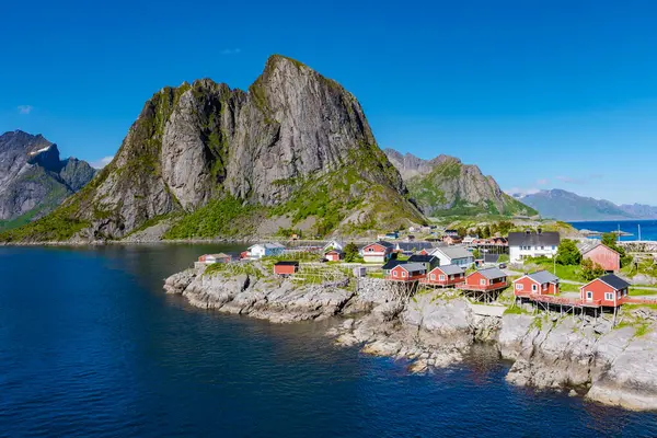 Norveç 'te fiyort boyunca yerleşmiş canlı kırmızı evlerle resim köyü. Uzun dağlar açık mavi gökyüzünün altında belirgin bir şekilde yükselir, Hamnoy, Lofoten, Norveç 'teki balıkçı köyü.