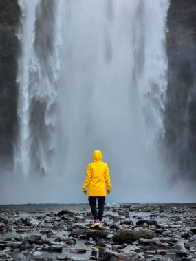 Bir gezgin, parlak sarı bir yağmurlukla İzlanda 'daki güçlü Skogafoss Şelalesine bakıyor. Sis onların etrafında yükseliyor ve bu çarpıcı doğal manzaraya bir merak duygusu ekliyor..