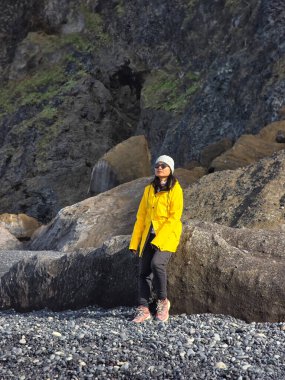 Sarı ceketli bir kadın Reynisfjara Sahili 'ndeki karanlık volkanik kayalara karşı duruyor. Okyanus dalgaları yavaşça çarpıp İzlanda 'nın eşsiz volkanik manzarasını aydınlatırken güneş gölgeleri salıyor..