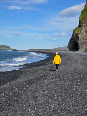 Parlak sarı ceketli yalnız bir gezgin, İzlanda 'daki geniş mavi gökyüzünün altında dramatik uçurumlar ve nazik dalgalarla çevrili Reynisfjara Sahili' nin siyah volkanik kumları boyunca yürür.