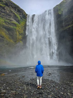 Bir gezgin İzlanda 'daki güçlü Skogafoss şelalesinin önünde huşu içinde duruyor, yemyeşil ve çağlayan suyun sisi ile çevrili. Atmosfer büyüleyici ve huzurlu..
