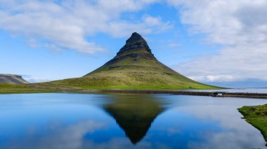 Kirkjufell Dağı, İzlanda 'nın altındaki sakin sularda kusursuz bir şekilde yansıtılan derin mavi gökyüzünün dingin arkaplanına karşı dimdik ve simgeli duruyor.