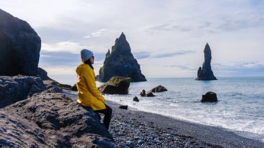Reynisfjara Sahili İzlanda 'nın kara kumlarında oturan bir gezgin bulutlu bir gökyüzünün altında okyanustan yükselen görkemli deniz yığınlarını seyrediyor. Sakin dalgalar yavaşça kıyıya vurur.