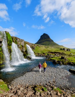 Kirkjufell şelalesi yakınlarındaki nehir kıyısında gezinen iki yolcu. Bereketli bitki örtüsü ve çarpıcı dağ manzaralarıyla çevrili bölge İzlanda 'nın doğal güzelliklerinden huzurlu bir kaçış sunuyor..