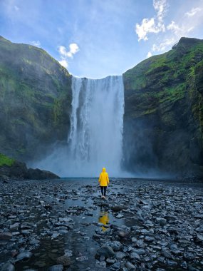 Skogafoss şelalesi İzlanda 'daki görkemli kayalıklardan aşağı akarak mistik bir atmosfer yaratır. Sarı yağmurluklu kadın şelalenin yanında duruyor. 