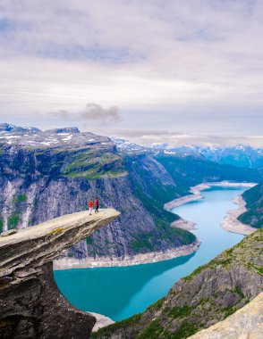 Maceraperest yürüyüşçüler, Norveç 'in dramatik Trolltunga uçurumunun kenarında dikiliyorlar. Aşağıdaki büyüleyici Lysefjord manzarasına hayret ediyorlar. Görkemli dağlar ve bulutlu bir gökyüzünün altında sakin sularla çevrili.