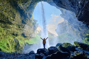 İzlanda 'daki Kvernufoss şelalesinin nefes kesici manzarası. Şelale çağlayan suyun altında yalnız bir gezginin huşu içinde durduğu yer. Güneş ışığı mağaranın içinden süzülüp bereketli manzarayı aydınlatıyor..