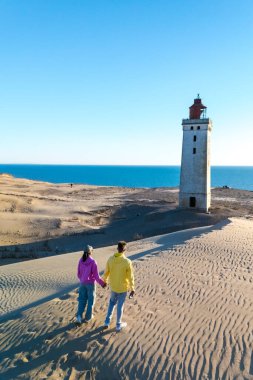 İki kişi el ele geziyor, yayılan kum tepeleri boyunca, tarihi bir deniz feneri ile mavi okyanus zeminine karşı görkemli bir şekilde duruyor. Rubjerg Knude Deniz feneri Danimarka Jutland