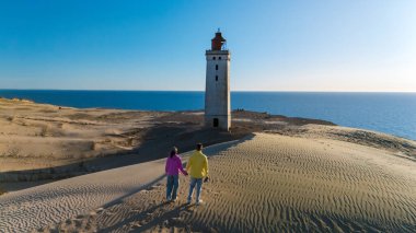 El değmemiş kumulların ve açık mavi gökyüzünün arkaplanının karşısında, bir çift el ele yürür simgesel Rubjerg Knude deniz fenerine doğru, Danimarka 'nın Kuzey Denizi' nde güzel bir günün tadını çıkarırlar.