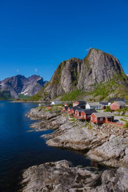 Lofoten Norveç 'in sakin sahil şeridini kayalık kıyılara kurulmuş geleneksel kırmızı kulübelerle deneyin. Açık mavi gökyüzünün altında nefes kesici dağlar ihtişamlı bir şekilde yükselir..