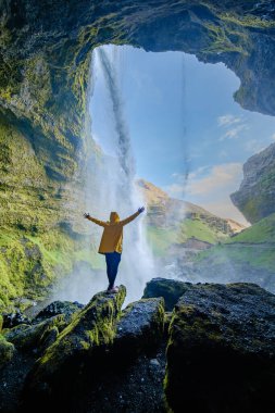 İzlanda 'daki çarpıcı Kvernufoss şelalesine hayranlıkla bakan bir gezgin, çağlayan suların altında durup İzlanda' nın doğal güzelliğinin tadını çıkarıyor.