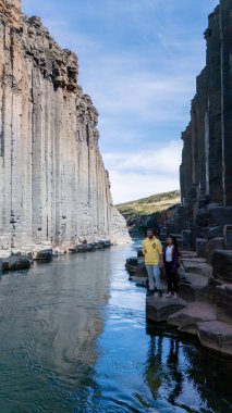 Studlagil Kanyonu 'ndaki kristal berrak nehrin yanında iki ziyaretçi duruyor. Etrafı bazalt kayalıklarla çevrili. Güneş ışığı görkemli manzarayı aydınlatır, macera için çarpıcı bir zemin yaratır..