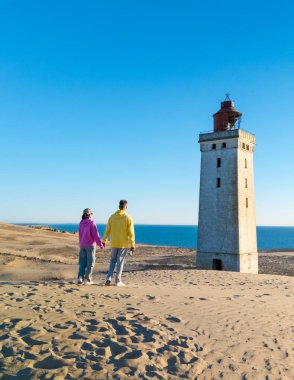 Bir çift, berrak mavi gökyüzüne karşı hayret verici Rubjerg Knude deniz fenerine hayran el ele durur. Danimarka 'nın kumulları resim gibi bir zemin oluşturuyor. Keşif ve romantizm için mükemmel..