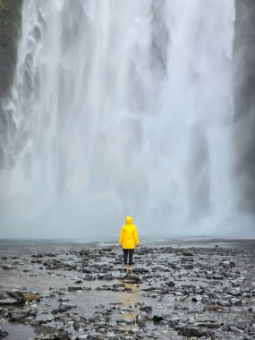 Parlak sarı bir yağmurluk giyen biri kayalık arazide duruyor, görkemli Skogafoss şelalesine bakıyor. Su arka planda dramatik bir şekilde yükselir ve sisli bir atmosfer yaratır..