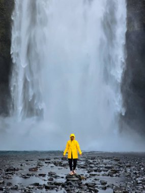 Skogafoss şelalesinin güçlü şelalelerinin ortasında parlak sarı yağmurluklu bir kadın İzlanda 'nın nefes kesici doğal güzelliğinin tadını çıkarıyor..
