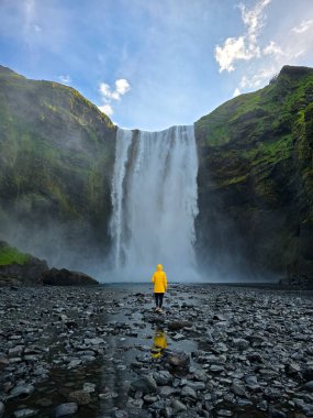 İzlanda 'daki Skogafoss şelalesinin hayranlık uyandırıcı manzarası. Parlak sarı yağmurluk giymiş bir gezgin, taşlı bir arazinin ortasında duruyor..
