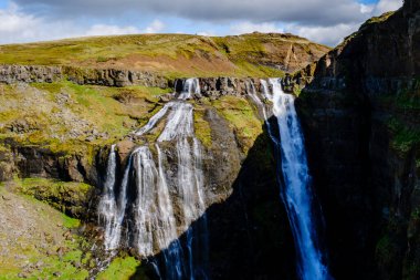 Güçlü bir şelale, yemyeşil ve İzlanda 'nın engebeli arazisiyle çevrili sarp kayalıklardan aşağı yuvarlanıyor. Mavi gökyüzünün altındaki dramatik manzara doğa meraklılarını ve fotoğrafçıları kendine çeker..