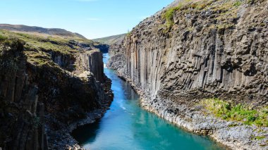 İzlanda 'da bulunan Studlagil Kanyonu' nda eşsiz bazalt sütunlar ve turkuaz sular sergileniyor. Ziyaretçiler, açık mavi gökyüzü altında doğa sanatına hayret ederek, manzaralı patikalarında yürüyüş yapmayı severler..