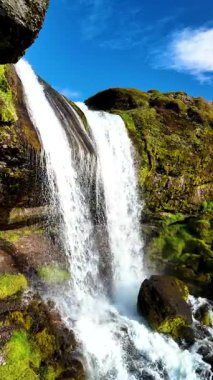Görkemli Selvallafoss şelalesi, canlı yeşil yosunlar ve eşsiz jeolojik oluşumlarla çevrili kayalık uçurumlardan aşağı yuvarlanıyor. Güneş ışığı huzurlu manzarayı aydınlatır., 