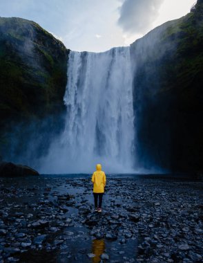 İzlanda 'daki Skogafoss şelalesinin nefes kesen güzelliğini deneyimleyin. Sarı yağmurluk giymiş bir ziyaretçi gün batımında düşen su tarafından büyülenir.
