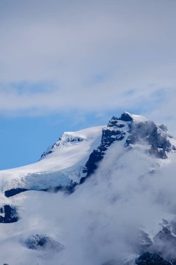 Bulutlarla kısmen örtülü Svinafellsjokull Buzulu 'nun nefes kesici güzelliğini deneyimleyin, berrak mavi gökyüzüne karşı çarpıcı buzlu zirvelerini sergiliyor. İzlanda doğasının gerçek mücevheri..
