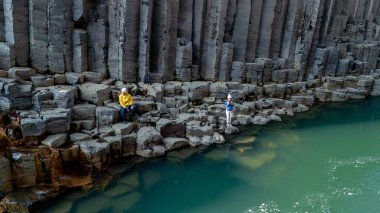 Su yavaşça Studlagil Kanyonu 'ndan akar, ziyaretçiler parlak bir gökyüzünün altında nehir yatağından yükselen etkileyici bazalt sütunları keşfeder.. 