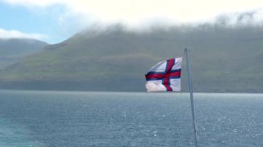İzlanda 'nın dingin güzelliğini hissedin. Faroe bayrağı sakin denizin üzerinde hafif bir esintide dalgalanıyor. Arkaplanda yemyeşil dağlar yükselir ve resim gibi bir ortam yaratır..