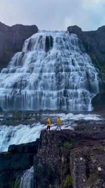 Ziyaretçiler, İzlanda 'nın kayalık manzarasına karşı şelalenin parladığı görkemli Dynjandi şelalesine hayranlıkla bakıyorlar..
