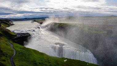 Soluk kesen Gullfoss şelalesi kayalık kayalıkların üzerinden İzlanda 'nın yemyeşil ve dramatik manzaralarıyla çevrili aşağıdaki dolambaçlı nehre dökülür.