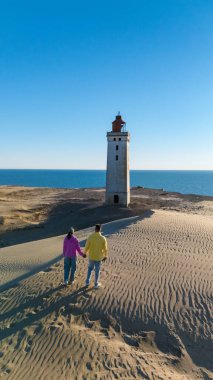 Kumlu sahil boyunca el ele yürür, denizin nefes kesici manzarasının ve Rubjerg Knude deniz feneri Kuzey Jutland 'da açık mavi gökyüzünün altında büyüleyici bir deniz fenerinin tadını çıkarır.