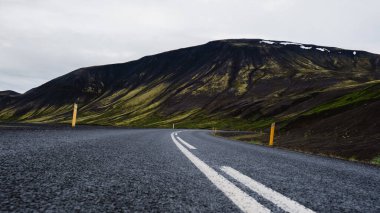 İzlanda 'nın engebeli arazileri boyunca uzanan ve kendine has dağlarla çevrili boş bir yol. Yeşillikler koyu volkanik kayalarla tezat oluşturuyor ve zarif bir doğal duvar halısı oluşturuyor..