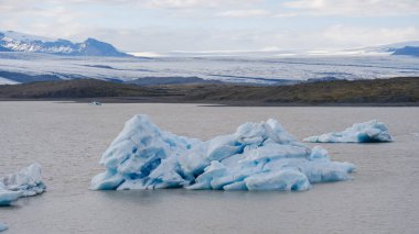 Işıl ışıl bir günde İzlanda 'daki bir buzul gölünün sakin suları boyunca sessiz mavi buzdağları sürükleniyor. Göz kamaştırıcı arka planda, yüksek buzullar ve engebeli dağlar görkemli doğalar sergiliyor..
