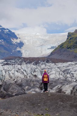 Yalnız bir yürüyüşçü kayalık arazide duruyor ve geniş Svinafellsjokull Buzulu 'na hayran kalıyor. Buzlu yüzey parlak bir gökyüzü altında parıldıyor, İzlanda 'nın doğal güzelliğini ve cazibesini vurguluyor..