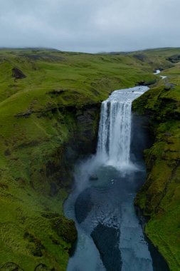 Skogafoss şelalesi, yeşil yeşilliklerle çevrili İzlanda 'daki yemyeşil uçurumların üzerinden güçlü bir şekilde akar. Sis, sular alçaldıkça yükselir ve hava kararırken dramatik manzaralar arasında sakin bir atmosfer yaratır..