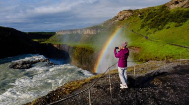 Bir gezgin Gullfoss şelalesinin kenarında duruyor, sislerin arasından yükselen canlı bir gökkuşağına bakıyor. Bereketli yeşil çevre büyülü atmosferi güçlendirir..