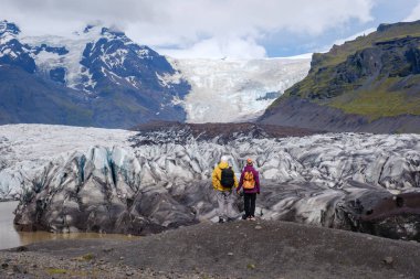 Svinafellsjokull Buzulu 'nun dramatik ve buzlu oluşumları arasında iki maceracı duruyor. Arka planda görkemli dağlar beliriyor, açık bir günde İzlanda 'nın engebeli güzelliğini gözler önüne seriyor..