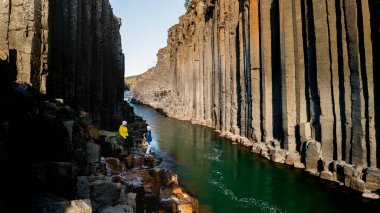 İzlanda 'daki Studlagil Kanyonu' nun göz kamaştırıcı güzelliğini keşfedin. Yüksek bazalt sütunlar ve sakin yeşil bir nehir. Ziyaretçiler güneşli bir günde doğal güzelliklere hayran kalırlar..