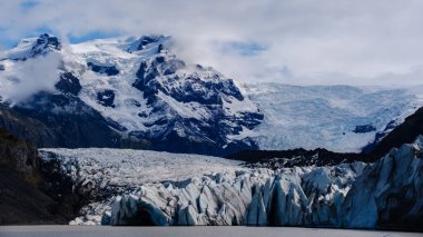 Svinafellsjokull Buzulu 'nun pırıl pırıl buz mavisi genişliğinde yükselen engebeli dağ zirveleri nefes kesici bir zemin oluşturur..