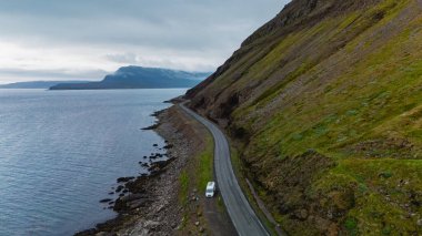 Bir karavan, İzlanda kıyı şeridinin sakin sularında yosun kaplı yüksek uçurumlarla çevrili dolambaçlı bir yolda ilerliyor. Batı fiyortları İzlanda