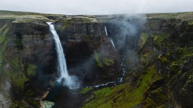 Haifoss şelaleleri engebeli kayalıklar ve sislerle çevrili canlı bir kanyona dökülüyor. Sahne, İzlanda 'nın bulutlu bir gökyüzü altında dramatik manzaralarını yakalıyor..