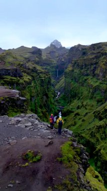 Görkemli dağlar, Mulagljufur Kanyonu 'nun yemyeşil alanının tepesinde iki maceraperest kayalıklarda durup çağlayan şelalelere bakıyor. Huzurlu manzara görkemini sergiliyor..
