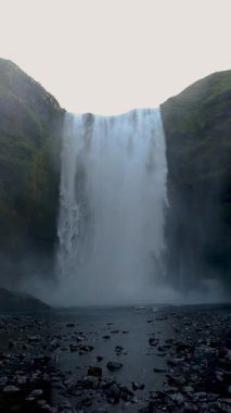 Skogafoss şelalesi, İzlanda 'nın göz kamaştırıcı doğal harikası, yosun kaplı uçurumların üzerinden zarif bir şekilde kayalık bir nehir yatağına yuvarlanıyor. Engebeli manzaranın büyüleyici güzelliğini tecrübe et..