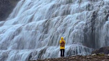 İzlanda 'nın, Dynjandi şelalelerini sersemleten şelalesini, parlak sarı ceketli bir yürüyüşçünün çağlayan suyun önünde duruşu gibi hissedin. Etrafı engebeli manzaralarla çevrili, yakınlarda büyüleyici bir güzellik beliriyor..