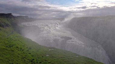 Gulfoss şelalesi kayalık kayalıklardan aşağı yuvarlanır. Etrafı yemyeşil çayırlar ve İzlanda 'daki sisli sularla çevrilidir.