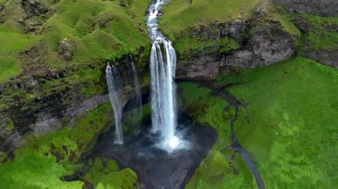 İzlanda 'daki Seljalandsfoss şelalesinin nefes kesici güzelliğini tecrübe edin. Su zarif bir şekilde yeşil kayalıklardan aşağı yuvarlanarak berrak gökyüzüne karşı çarpıcı bir doğal görüntü oluşturur..
