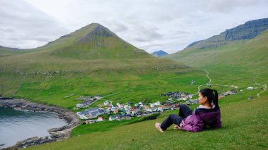Bereketli yeşil bir manzaraya yerleşmiş genç bir Asyalı kadın Faroe Adaları 'ndaki Gjogv köyünün huzurlu güzelliğinin tadını çıkarıyor..