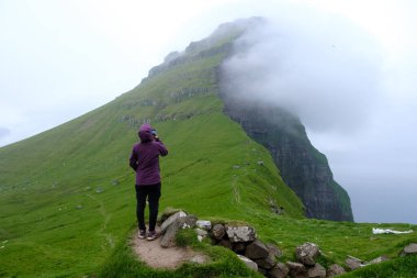 Bir yürüyüşçü yemyeşil bir yolda duruyor ve Faroe Adaları 'nın dramatik uçurumlarına bakıyor. Sis dağların üzerinde yuvarlanarak Kallur Deniz Feneri 'nde mistik bir atmosfer yaratıyor.