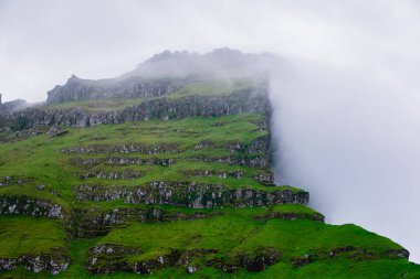 Faroe Adaları 'nda sis ve bulutlarla kaplı engebeli uçurumlar kulesi. Dramatik manzara doğanın saf güzelliğini ve dingin atmosferini gözler önüne seriyor, Kallur Deniz Feneri, Kalsoy Adası
