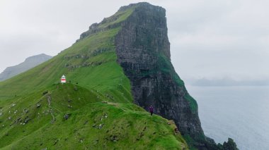 Faroe Adaları 'ndaki Atlantik Okyanusu' ndan dramatik uçurumlar yükseliyor, canlı yeşil manzaralar ve sakin bir deniz feneri sergiliyorlar. Kaşifler engebeli arazide geziniyorlar, Kallur Deniz feneri Kalsoy. 